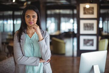 Asian woman standing with hand touching chin in office with computer monitor and glass partitions