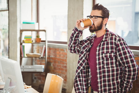 Man standing at loft office desk holding disposable coffee cup beside computer monitor, copy space