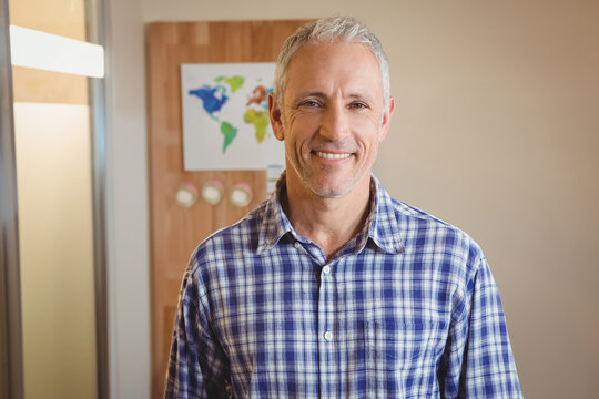 Male in plaid shirt standing and smiling in office against wood-paneled wall with map and clocks