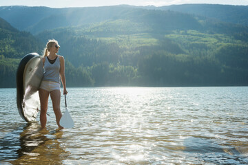 Woman wading with paddleboard in clear lake wearing sunglasses and standing before sunlit mountains
