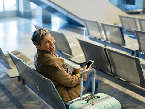 Senior woman sitting airport chair beside mint green suitcase holding coffee cup, phone, copy space