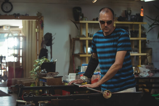 Mature male shaping molten glass orb on glassblowing pipe at metal bench in workshop with tools - Powered by Adobe