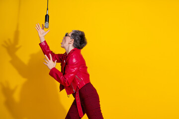 Stylish woman with a microphone against a yellow background showing enthusiasm wearing a trendy red leather jacket