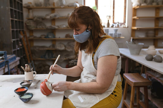 Female potter wearing mask and painting red glaze onto ceramic bowl at worktable in pottery studio - Powered by Adobe