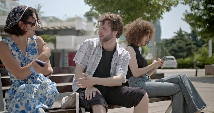 Three friends sit on a bench, engrossed in their phones, ignoring each other. One friend tries to engage in conversation but is ignored.