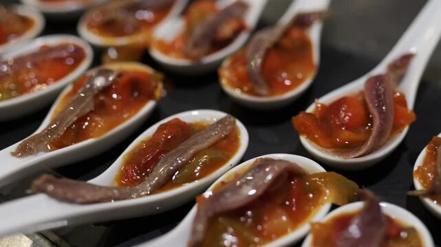 Contemporary ceramic spoons featuring ratatouille garnished with anchovies, resting on dark background with refined culinary presentation