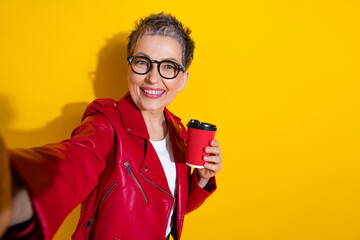 Stylish senior woman in red leather jacket with coffee cup on yellow background promoting fashion and lifestyle concepts