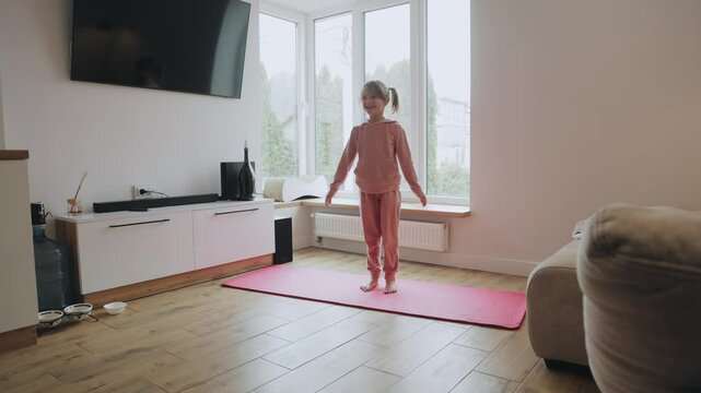 Young girl doing jumping jacks on yoga mat in living room. Happy child performs jumping jacks exercise on a pink yoga mat at home, promoting healthy lifestyle and fitness from a young age.