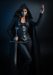portrait of mysterious hooded woman in black cloak, gothic fantasy or dystopian character, holding sword weapon. lit with moody cinematic shadows, studio scene on dark isolated background