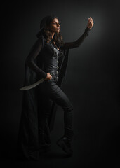 portrait of mysterious hooded woman in black cloak, gothic fantasy or dystopian character, holding sword weapon. lit with moody cinematic shadows, studio scene on dark isolated background