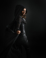 Naklejka premium portrait of mysterious hooded woman in black cloak, gothic fantasy or dystopian character. posing with gestural magical hand poses. lit with moody shadows, studio scene on dark isolated background