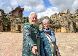 A smiling elderly couple takes a selfie during a sightseeing trip to an archaeological site,...