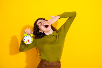 Sleepy girl holding a colorful alarm clock in a bright yellow background, wearing a green shirt