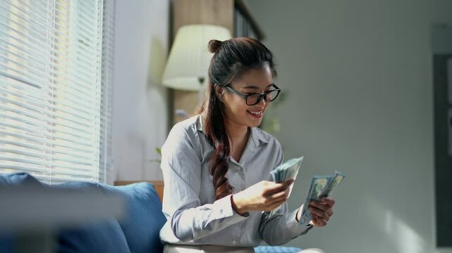 Young professional businesswoman sitting comfortably on home sofa, counting american dollars with confident smile, representing financial success and wealth