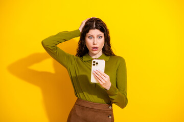 Surprised young woman in a green top holding a smartphone against a yellow background expressing...
