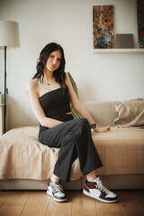 A young woman sits comfortably on a beige couch in a stylish living room. She is wearing a black jumpsuit paired with sporty sneakers, enjoying a serene moment