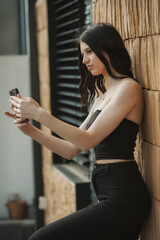 A young woman with long dark hair, dressed in a black crop top and pants, is sitting against a textured wall while focused on her smartphone