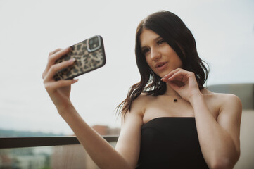 A woman with long dark hair enjoys taking a selfie on a balcony. She is dressed in a stylish outfit and poses playfully with a city skyline in the background