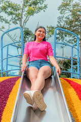 Young Latina woman enjoying a fun moment sliding down in a colorful playground under a sunny sky
