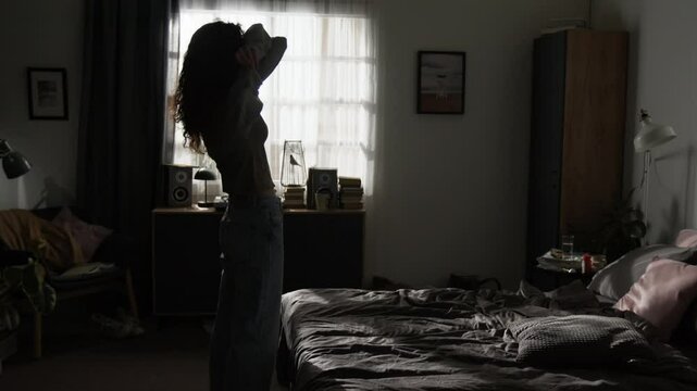 Depressed young woman taking off her sweater and laying down on bed, covering her face in dark sunlit bedroom