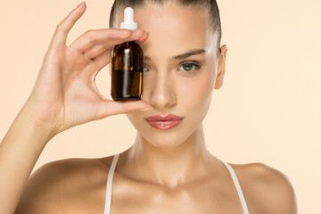 Fototapeta premium Beautiful young woman holding amber glass dropper bottle near face, showcasing skincare product on beige background.
