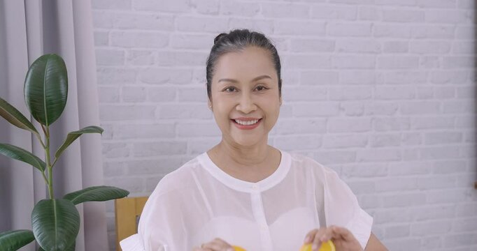 Asian senior female smiling brightly while holding two halves of fresh orange near face, symbolizing natural skincare, healthy lifestyle, beauty vitamins and joyful aging in playful moment