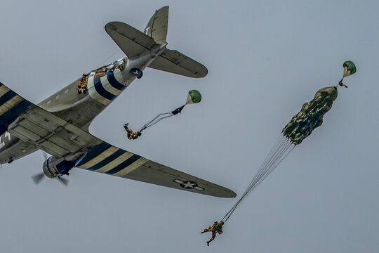Douglas C-47 with D-Day invasion stripes releases paratroopers and cargo during a WWII-style reenactment flight under an overcast sky.