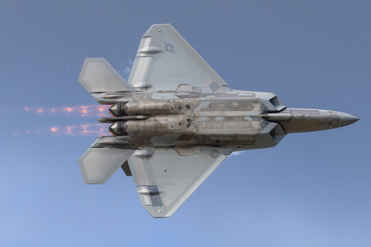 U.S. Air Force F-22 Raptor rockets past with blazing afterburners, displaying its angular stealth profile and twin-vectoring nozzles against a clear blue sky during an airshow flyby.