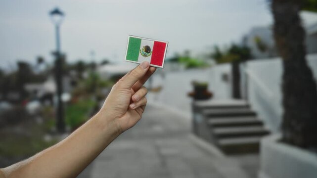 Man holding a small mexican flag embroidered patch with one hand, standing outdoors on a street in a town, capturing a sense of cultural pride and national identity.
