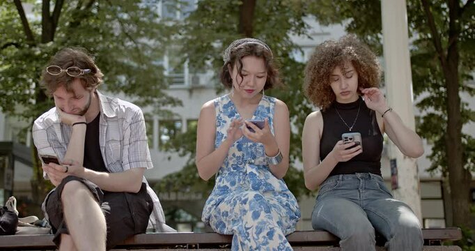Three friends sit together on a bench, engrossed in their phones, ignoring each other and the world around them.