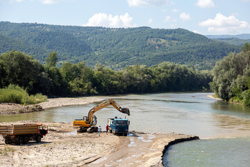 An excavator and dump trucks extract gravel from a riverbed, with lush green mountains and forests in the background on a sunny day.