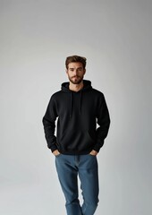Relaxed young man wearing a black hoodie and jeans, posing confidently with hands in pockets against a neutral studio backdrop