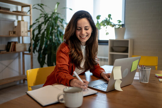 Smiling freelancer taking notes while working on laptop at home office