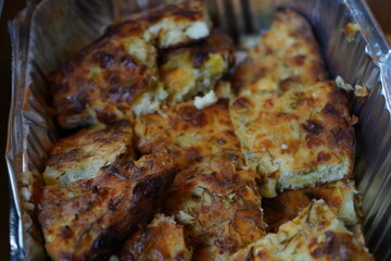 Homemade zucchini and cheese pie with herbs - Greek cuisine, cretan hospitality and Mediterannean diet - Golden-brown pie, chopped into rectangular pieces, in an aluminum tray. 