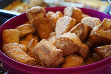 Golden-brown pastries fill a purple bowl, showcasing their flaky texture. These bite-sized, puff pastry treats are neatly arranged, highlighting their crisp, layered surface. 