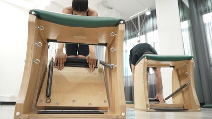 Clients practicing a standing stretch using Wunda Chair equipment during a Pilates workout for improving flexibility, leg strength at gym. Attractive group stretching and bending down. Habituate.