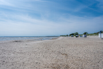 A serene sandy beach in Parnu, Estonia, features calm waters, a clear blue sky, scattered beach huts, sparse greenery, and low dunes in the background.