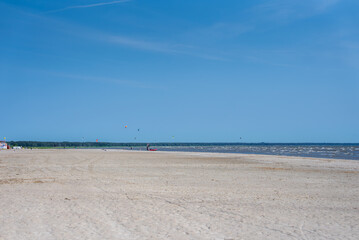 A tranquil beach in Parnu, Estonia, featuring calm waters, a clear blue sky, colorful kites in the distance, and a small structure on the left.