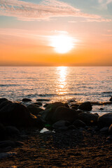 A serene sunset over the Baltic Sea in Parnu, Estonia, with warm orange hues, calm water reflections, a rocky shoreline, and wispy clouds in the sky.
