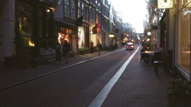 Blurred evening scene of a cyclist riding through a busy european city street with people walking and dining outdoors amidst illuminated shopfronts and cobblestone paths.