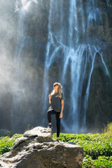 Obraz premium Young woman standing on a rock near big majestic waterfall at sunset in summer. Plitvice Lakes national park, Croatia. Lifestyle. Sporty girl on the stone near amazing waterfall. Hiking and trekking