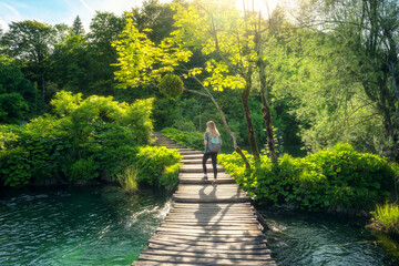 Young woman with backpack walking along a wooden path near stunning waterfalls, green trees in...
