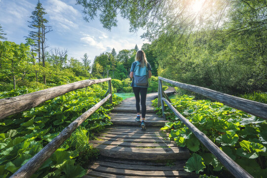 Young woman with backpack walking along a wooden path near stunning waterfalls, green trees in Plitvice Lakes national park, Croatia at sunset in summer. Female tourist. Sporty girl in forest. Hiking