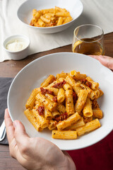 Vegan pasta with dried tomato served on a white deep plate by a hand, viewed from a 45 degree angle. Next to the dish sits a fork with a grey tissue, a glass of white wine, a small bolw with parmesano
