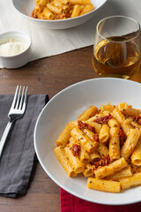 Vegan pasta with dried tomato served on a white deep plate, viewed from a 45 degree angle. Next to the dish sits a fork with a grey tissue, a glass of white wine, a small bolw with parmesano and the h
