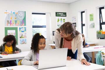 Female teacher helping girl using laptop in classroom, promoting learning and engagement, at school