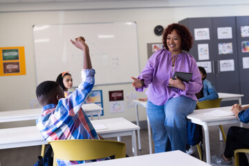 Female teacher engaging with diverse students in class, boy raising hand to participate, at school