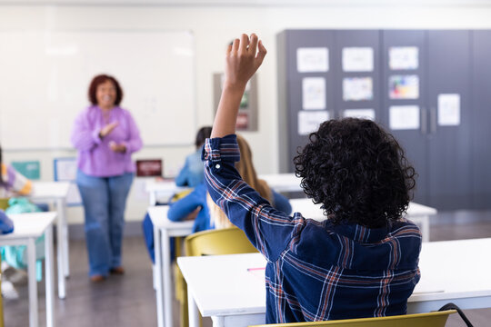Student raising hand in classroom while female teacher engaging with diverse students, at school