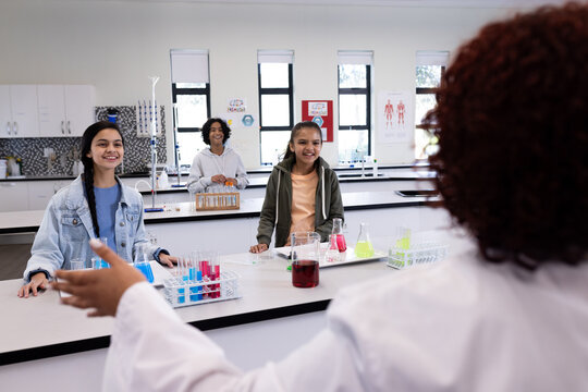 Diverse students in science class engaging with female teacher, surrounded by test tubes, at school