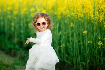  A little girl in a white dress and cat-shaped pink sunglasses stands by a bright yellow flower...